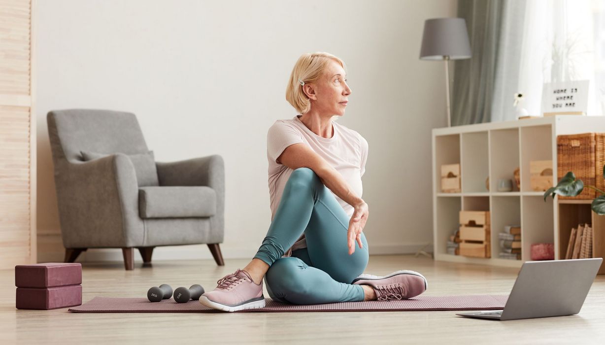 Ältere Frau macht Yoga zu Hause vor dem Rechner.