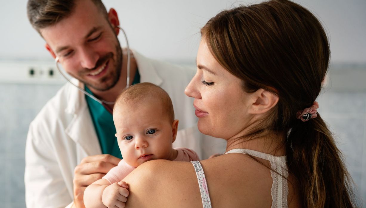 Baby blickt über die Schulter der Mutter, ein junger Kindarzt hört den die Lunge des Babys am Rücken ab.