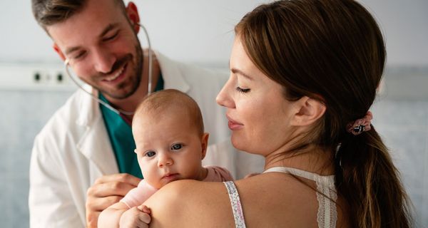 Baby blickt über die Schulter der Mutter, ein junger Kindarzt hört den die Lunge des Babys am Rücken ab.