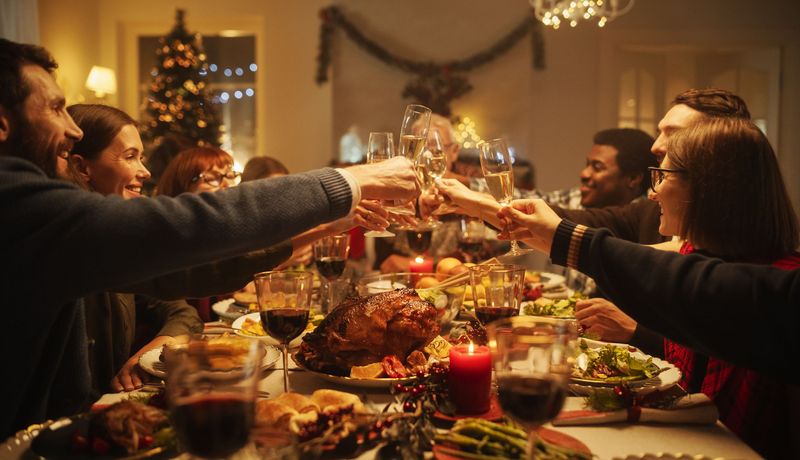 Eine große Gruppe sitzt um einen Weihnachtstisch herum. Viel Essen steht auf dem Tisch und alle stoßen mit Weingläsern an.