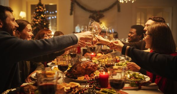 Eine große Gruppe sitzt um einen Weihnachtstisch herum. Viel Essen steht auf dem Tisch und alle stoßen mit Weingläsern an.