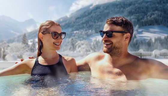 Ein Mann und eine Frau sitzen lächelnd in einem Whirlpool (Hot Tub), im Hintergrund sind verschneite Berge zu sehen.