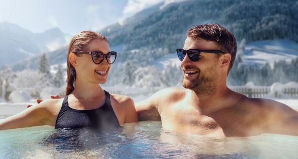 Ein Mann und eine Frau sitzen lächelnd in einem Whirlpool (Hot Tub), im Hintergrund sind verschneite Berge zu sehen.