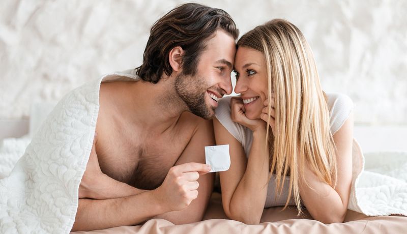 Nackter Mann und Frau liegen zusammen unter einer dünnen Decke. Sie lächeln sich an, der Mann hat ein Kondom in Verpackung in der Hand.