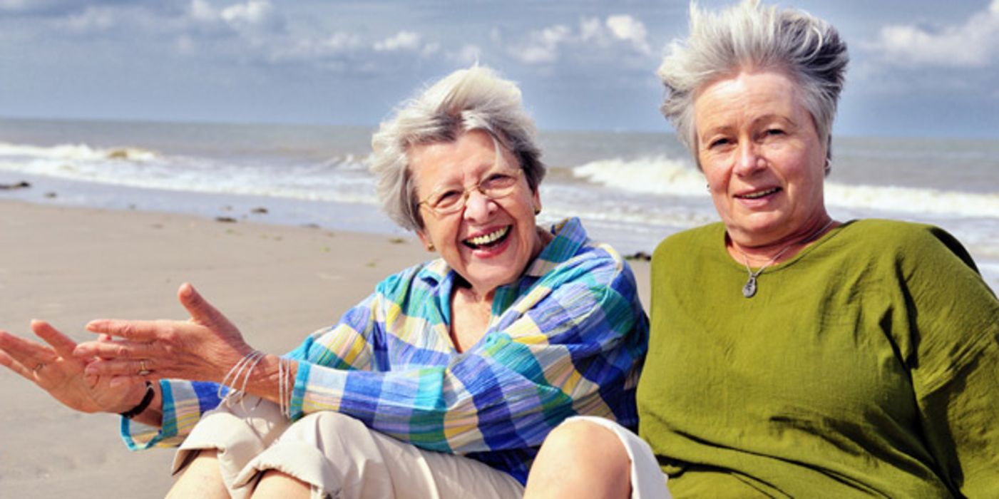 Zwei aktive Seniorinnen sitzen gut gelaunt am Strand und lassen sich den Wind durch die Haare wehen