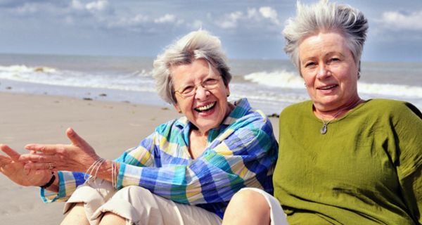 Zwei aktive Seniorinnen sitzen gut gelaunt am Strand und lassen sich den Wind durch die Haare wehen