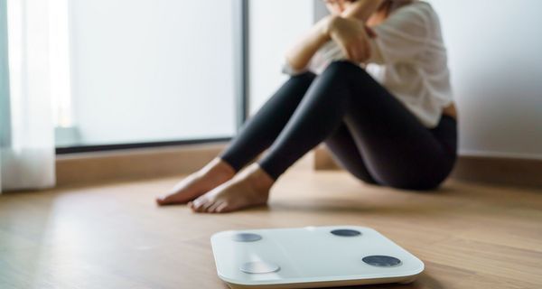 Im Vordergrund steht eine Waage, dahinter sitzt eine junge Frau am Boden und verbirgt das Gesicht in den Armen.
