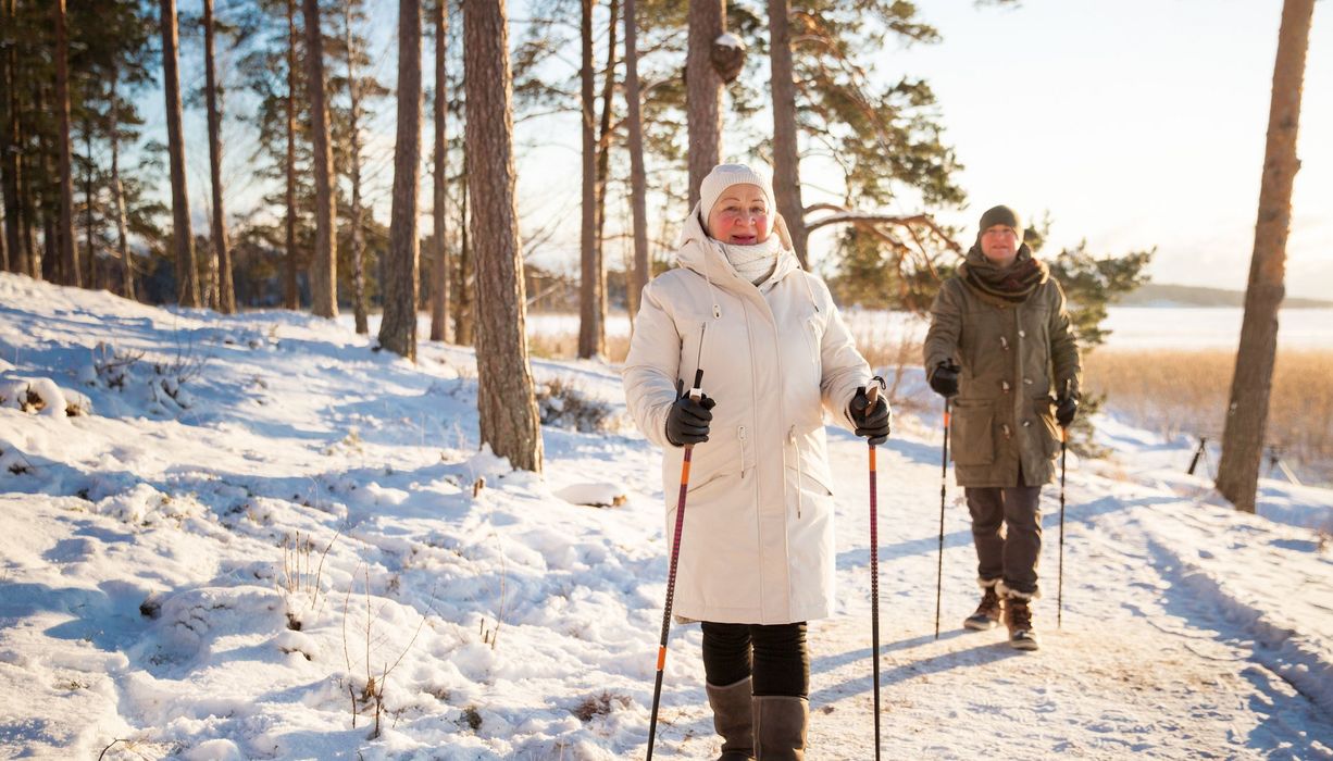 Eine ältere Dame und ein älterer Herr gehen in einem winterlichen Wald spazieren.