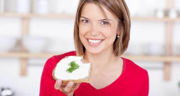 Küchenszene (im Hintergrund schemenhaft zu erkennen), Frau ca. 30, roter Pullover mit V-Ausschnitt, lächelt in die Kamera, in der linken Hand ein Butterbrot mit Kressegarnitur