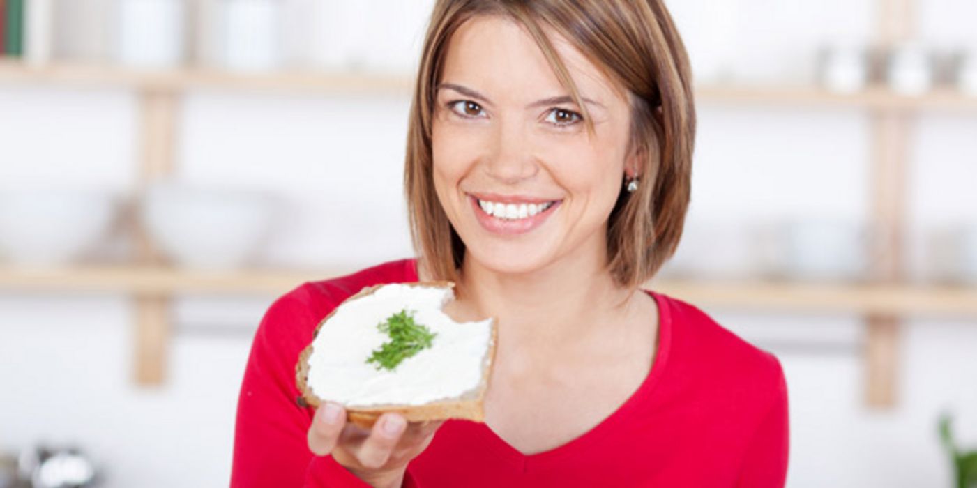 Küchenszene (im Hintergrund schemenhaft zu erkennen), Frau ca. 30, roter Pullover mit V-Ausschnitt, lächelt in die Kamera, in der linken Hand ein Butterbrot mit Kressegarnitur