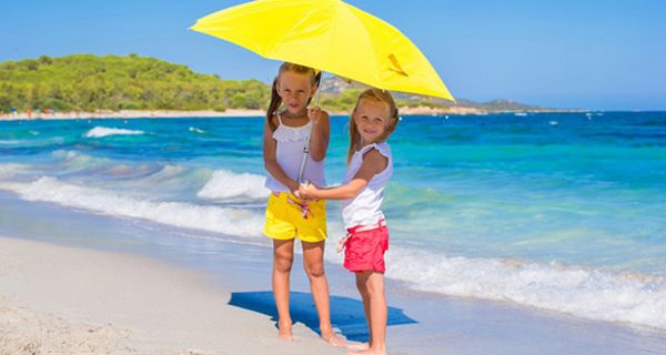 2 blonde Mädchen (ca. 9 und 6 Jahre), weiße Shirts, Shorts gelb bzw. blau, unter großem gelbem Sonnenschirm an weißem Strand am blauen Meer