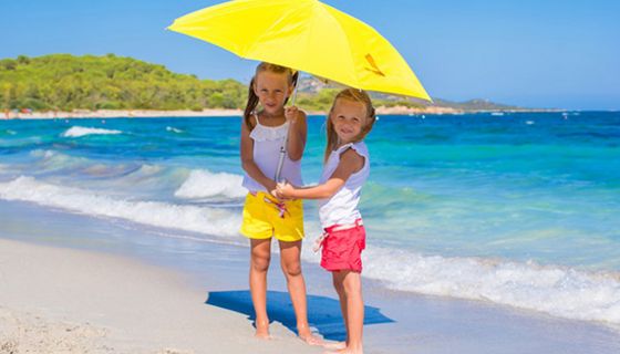 2 blonde Mädchen (ca. 9 und 6 Jahre), weiße Shirts, Shorts gelb bzw. blau, unter großem gelbem Sonnenschirm an weißem Strand am blauen Meer