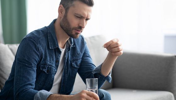 Mann blickt zweifelnd auf eine Tablette in seiner Hand. In der anderen hält er ein Glas Wasser.