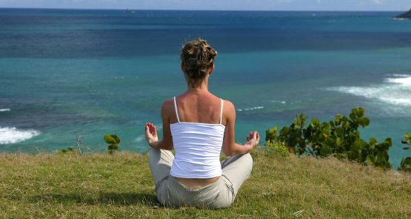 Frau von hinten in Meditationshaltung auf einer Wiese mit Blick aufs tiefer gelegene Meer