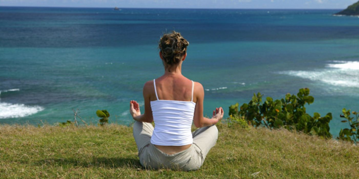 Frau von hinten in Meditationshaltung auf einer Wiese mit Blick aufs tiefer gelegene Meer