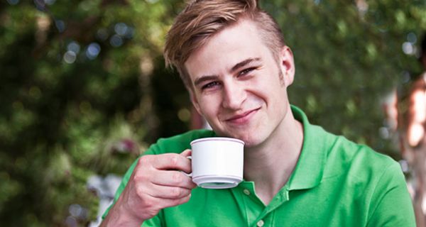 Frontalfoto: junger Mann in den 20ern, grünes Poloshirt, Kaffeetasse in der rechten Hand, freundlich lächelnd