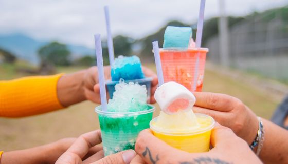 Vier Personen - nur ihre Hände sind zu sehen - stoßen mit bunten Slush-Getränken an. 