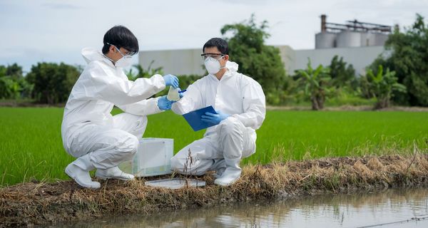 Zwei Männer in weißer Schutzkleidung und Masken hocken neben einem Fluss. Sie entnehmen mit Gefäßen Wasser aus dem Fluss.