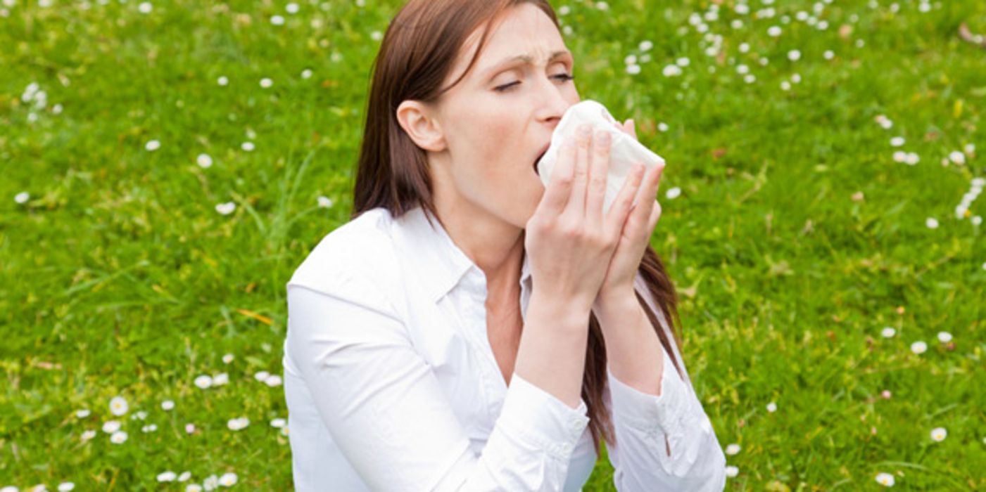 Vordergrund Frau im weißen Langarmshirt, braune Haare, niest in ein Taschentuch; im Hintergrund Wiese mit weißen Blümchen