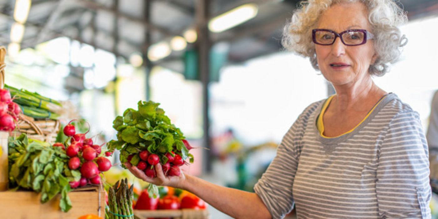 Moderne Seniorin beim Gemüsekauf in Markthalle: graue Locken, kinnlang, moderne Hornbrille, grau gestreiftes Shirt, gelb eingefasst, gelbe Uhr. Sie schaut in die Kamera