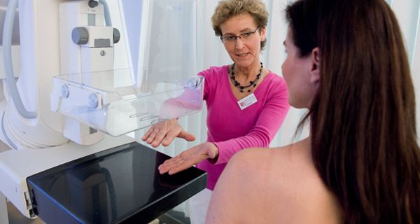 Mammografieszene, zu untersuchende Frau (lange, dunkle Haare) mit dem Rücken zur Kamera, etwas Profil zu sehen, Assitentin erklärt den Vorgang