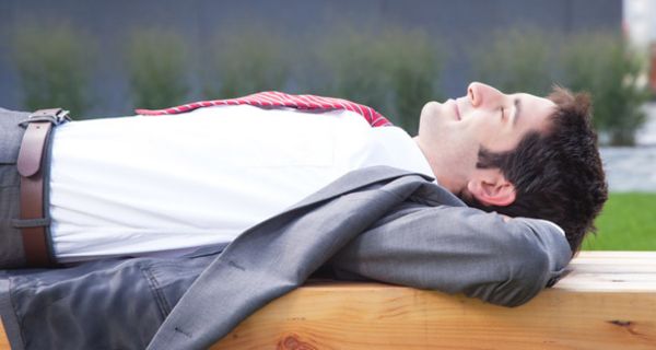 Junger Mann in Anzug und Krawatte liegt auf einer Bank und hat die Augen geschlossen.