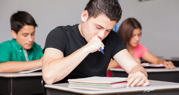 Ein Jugendlicher brütet in der Schulklasse über seinen Aufgaben, er hält den Stift in einer Faust nahe am Mund.. Im Hintergrund sind ein Mitschüler und eine Mitschülerin zu sehen. 