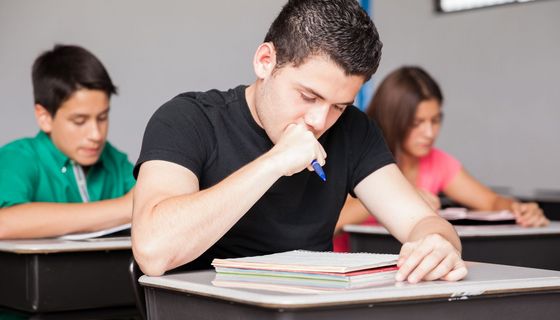 Ein Jugendlicher brütet in der Schulklasse über seinen Aufgaben, er hält den Stift in einer Faust nahe am Mund.. Im Hintergrund sind ein Mitschüler und eine Mitschülerin zu sehen. 