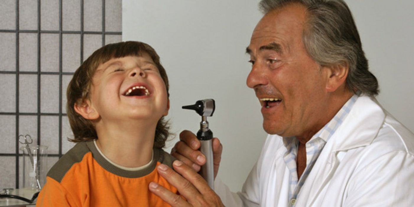 Szene beim Ohrenarzt: Älterer Arzt untersucht linkes Ohr eines Jungen (ca. 6 Jahre) und scherzt dabei mit diesem. Beide lachen.