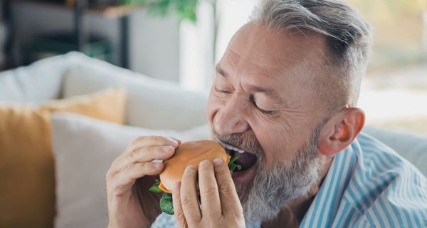 Mann zwischen 50 und 80 Jahren – er hat graue haare und einen grauen Bart – beißt in einen Burger.