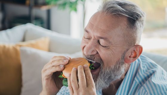 Mann zwischen 50 und 80 Jahren – er hat graue haare und einen grauen Bart – beißt in einen Burger.