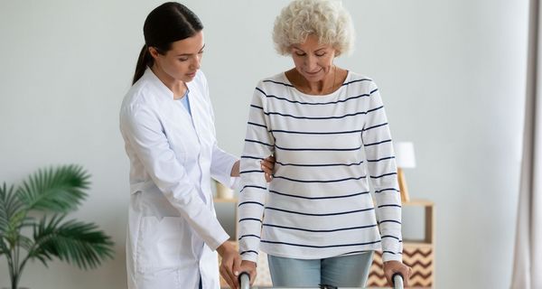 Eine ältere Dame stützt sich auf einer Gehilfe ab. Eine junge Frau im Kittel hält begleitend ihren Arm.