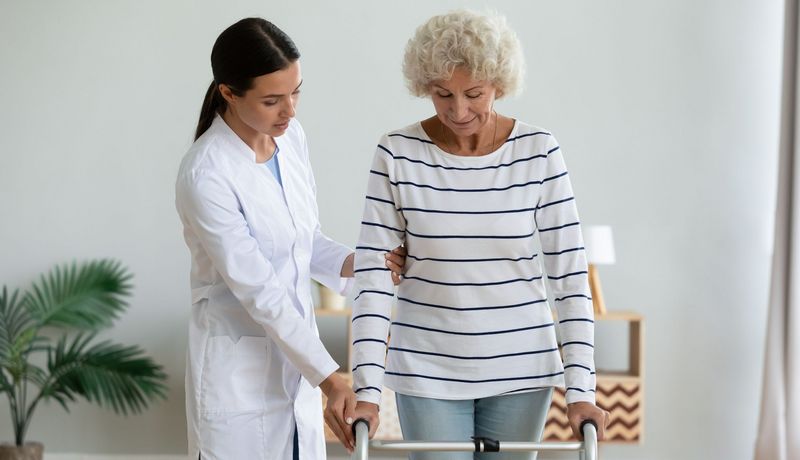 Eine ältere Dame stützt sich auf einer Gehilfe ab. Eine junge Frau im Kittel hält begleitend ihren Arm.