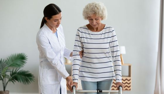 Eine ältere Dame stützt sich auf einer Gehilfe ab. Eine junge Frau im Kittel hält begleitend ihren Arm.