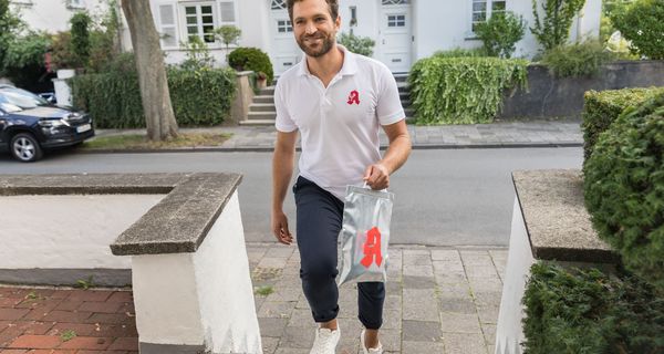 Mann mit Apothekenzeichen auf dem T-Shirt geht mit einer Arzneimitteltüte auf einen Hauseingang zu.