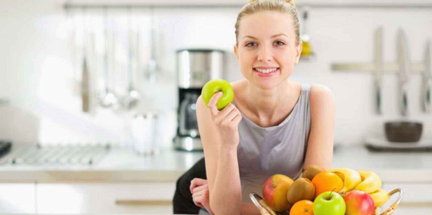 Junge in die Kamera lächelnde Frau mit blondem Dutt lehnt auf einem Küchentresen; vor sich einen Obstkorb, in der rechten Hand einen grünen Apfel.