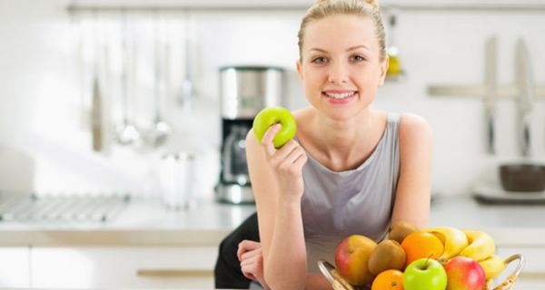 Junge in die Kamera lächelnde Frau mit blondem Dutt lehnt auf einem Küchentresen; vor sich einen Obstkorb, in der rechten Hand einen grünen Apfel.