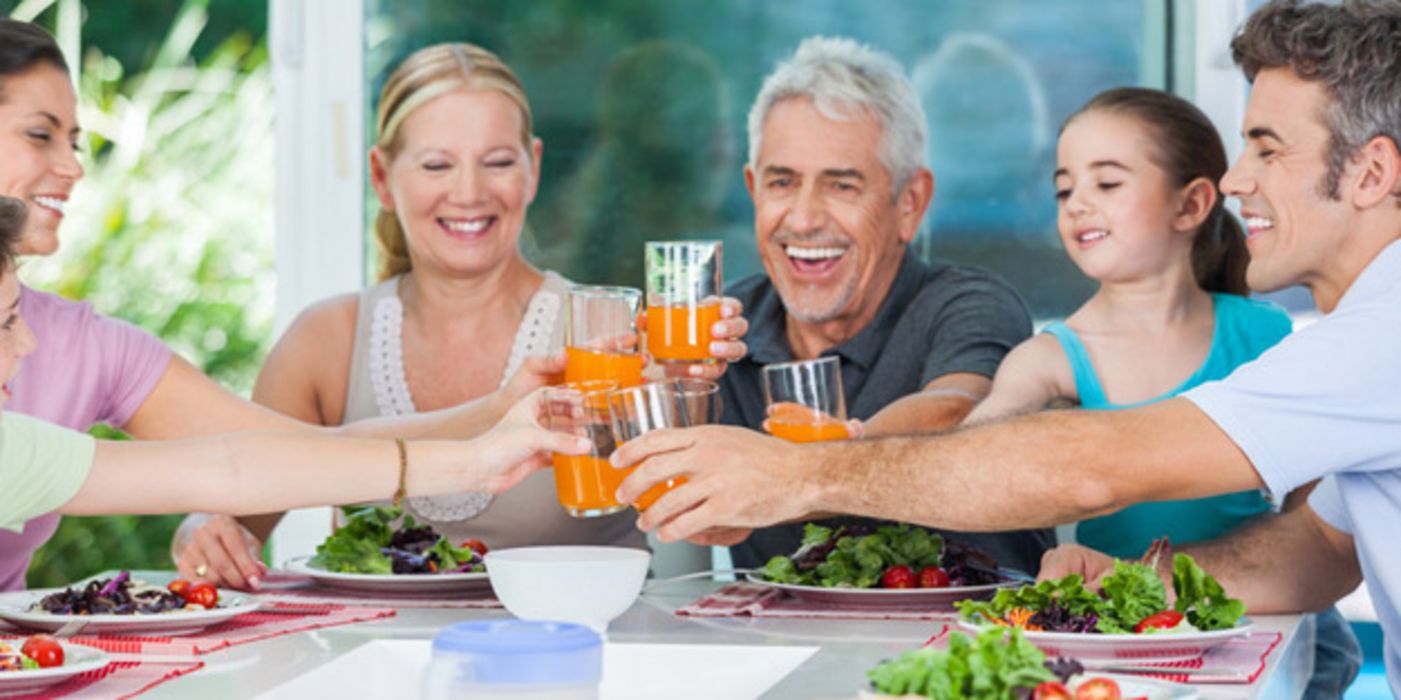 Fröhliche Familie, 3 Generationen, am Esstisch, Salate vor sich, stoßen mit Orangen-Limo an