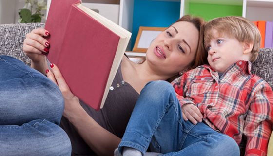 Mutter und Sohn (ca. 4jährig), beide leger in Jeans und Pulli bzw. Hemd gemütlich auf Couch gekuschelt. Mutter liest Sohn aus rotgebundenem großen Buch vor
