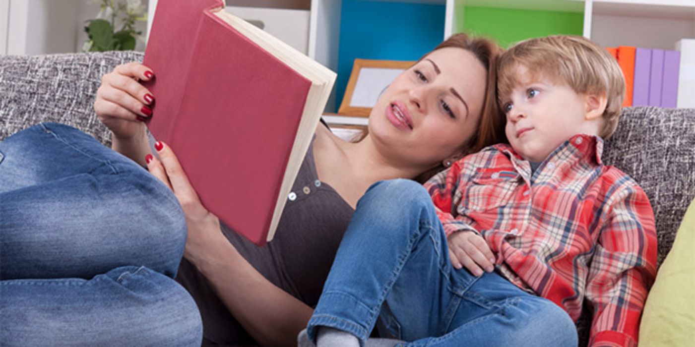 Mutter und Sohn (ca. 4jährig), beide leger in Jeans und Pulli bzw. Hemd gemütlich auf Couch gekuschelt. Mutter liest Sohn aus rotgebundenem großen Buch vor