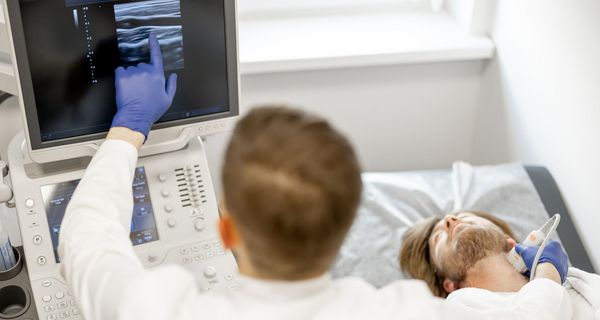 En Mann liegt auf einer Liege. Der Arzt neben ihm untersucht seine Halsschlagader mit einem Ultraschallgerät und deutet auf den Bildschirm.
