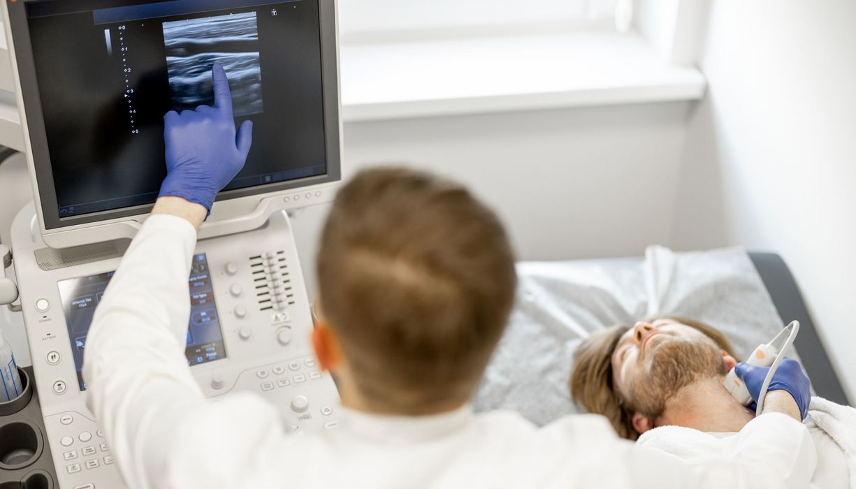 En Mann liegt auf einer Liege. Der Arzt neben ihm untersucht seine Halsschlagader mit einem Ultraschallgerät und deutet auf den Bildschirm.