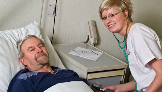 Mann um die 60 im Krankenhausbett, jüngere  Krankenschwester, die ihm den Blutdruck misst. Beide lächeln in die Kamera