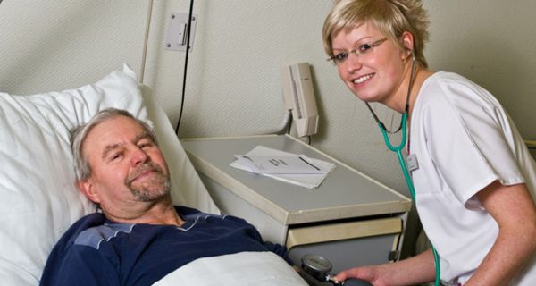 Mann um die 60 im Krankenhausbett, jüngere Krankenschwester, die ihm den Blutdruck misst. Beide lächeln in die Kamera