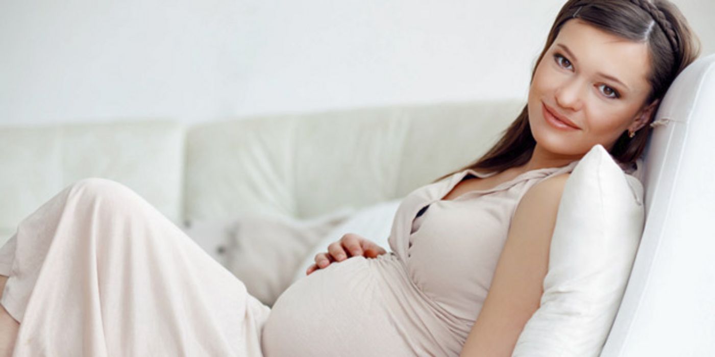 Schwangere, dunkle Haare, in hellem Hauskleid ohne Ärmel auf Couch, eine Hand auf Bauch, lächelnd in die Kamera schauend