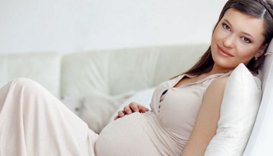 Schwangere, dunkle Haare, in hellem Hauskleid ohne Ärmel auf Couch, eine Hand auf Bauch, lächelnd in die Kamera schauend