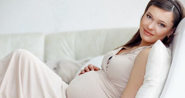 Schwangere, dunkle Haare, in hellem Hauskleid ohne Ärmel auf Couch, eine Hand auf Bauch, lächelnd in die Kamera schauend