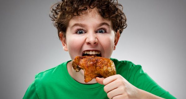 Frontalfoto eines Jungen mit dunklen Locken und grünem T-Shirt, der mit aufgerissenen Augen an einer Hühnerkeule nagt
