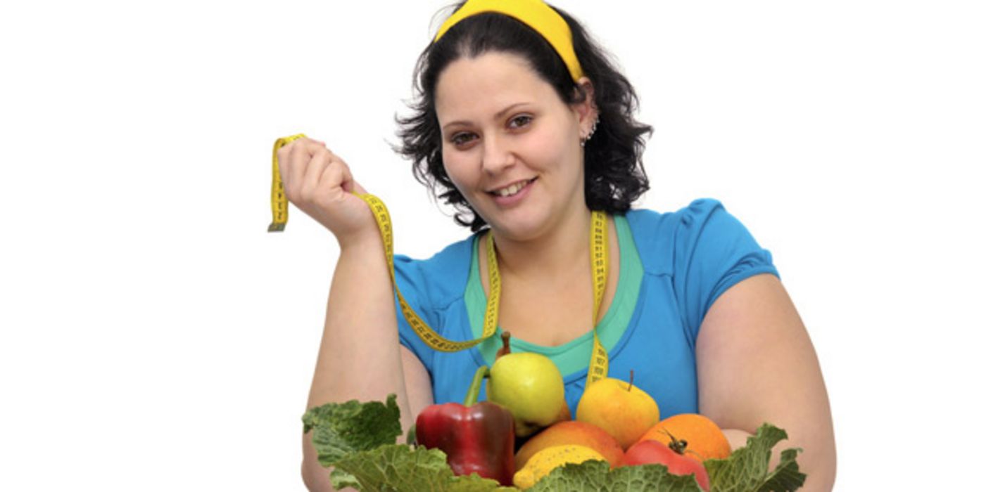 Übergewichtige, junge Frau mit Obst und Gemüse vor sich auf einem Tisch und einem Maßband um den Hals
