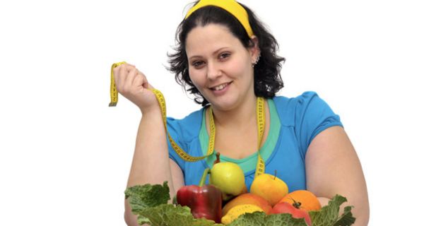 Übergewichtige, junge Frau mit Obst und Gemüse vor sich auf einem Tisch und einem Maßband um den Hals